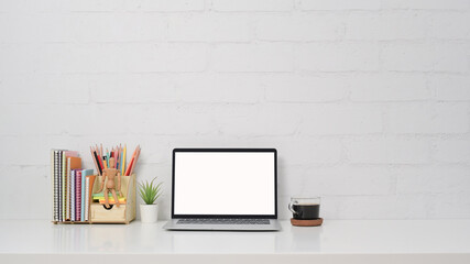 Front view computer laptop, coffee cup and stationery on white table against brick wall.