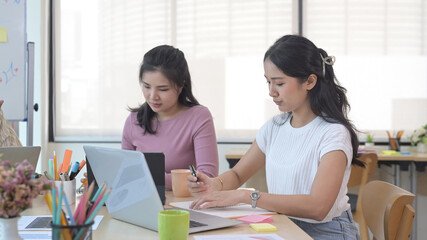 Two young creative women in casual clothes sitting at modern office and working on project together.