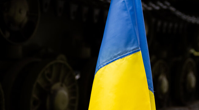 National flag of Ukraine, with battle tank in background. Protest manifestation against Russia invasion, war in Ukraine. Ukrainian banner, anti-war demonstration next to military armored vehicle.