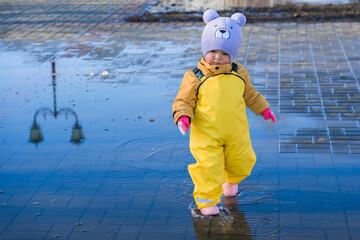 A child aged 2-3 years old dressed in a yellow rubber jumpsuit walks through puddles in the park