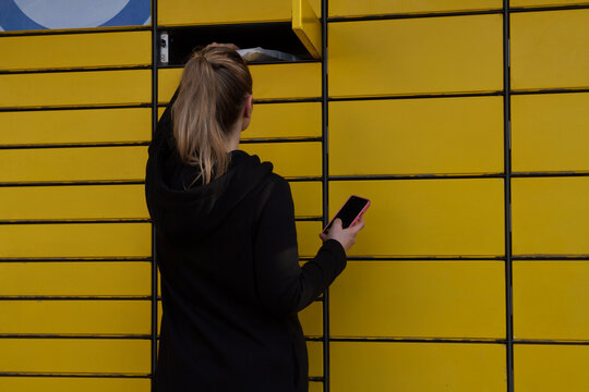 Beautiful woman picking up a package from a smart electronic steel parcel locker box, automatic mailboxes. Paczkomat delivery service, collection machine. Using app on her phone to open the door.