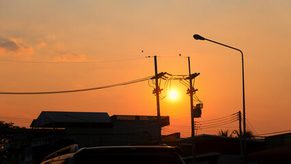 Silhouettes buildings, high voltage poles, streetlight poles, power lines and birds flying in the golden sky during sunrise or sunset.