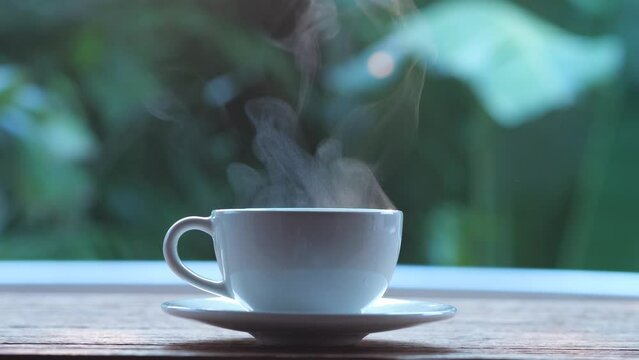 Hot coffee in the morning sunrise, soft steam smoke floated above the white coffee cup on the wooden table, put on a saucer and left hand grip. Hot coffee concept.