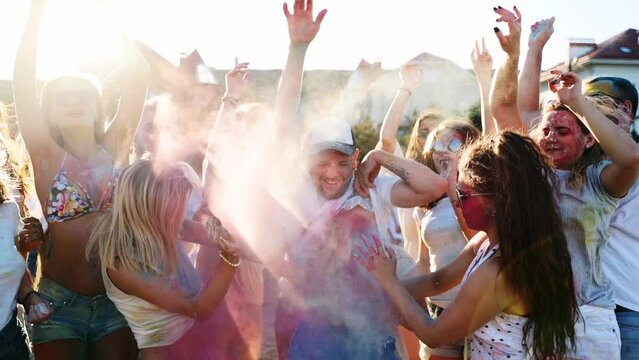 Young man surrounded by crowd gets splashes of colored powder onto belly by two women in slow motion. Friends have fun at hindu holiday of spring, colors, love. End of covid pandemic isolation.