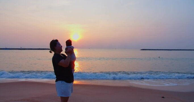 happiness Asian family mother is keeping on her arms holding playing and kissing cute adorable newborn baby infant girl on a seaside beach at sunrise morning during holiday vacation in Thailand