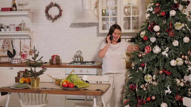 Happy smiling pregnant woman talking on the phone and decorating the Christmas tree in the modern loft kitchen.