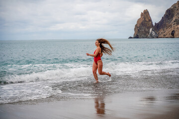 Fototapeta premium A beautiful and sexy brunette in a red swimsuit on a pebble beach, Running along the shore in the foam of the waves