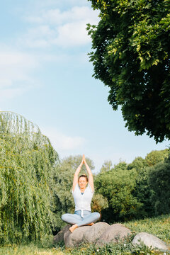 Young woman practicing yoga asana parvatasana sitting in lotus position on stone in nature. Sportswoman meditating exercising outdoors on sunny morning. Active healthy lifestyle