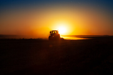 Tractor in sunset, farmer ploughs a field with tractor in sunset.  The tractor is backlit by the setting sun.  Day is turning night in the field. Beautiful agriculture concept idea photo. © Designerant