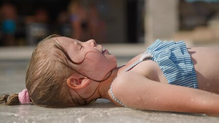 Cute blonde child in blue swimsuit plays splashing clear water lying on river shallow bottom at resort on sunny summer day close side view slow motion