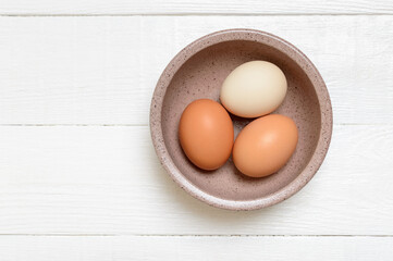 Three chicken farm eggs in a bowl on a white wooden background. Top view.