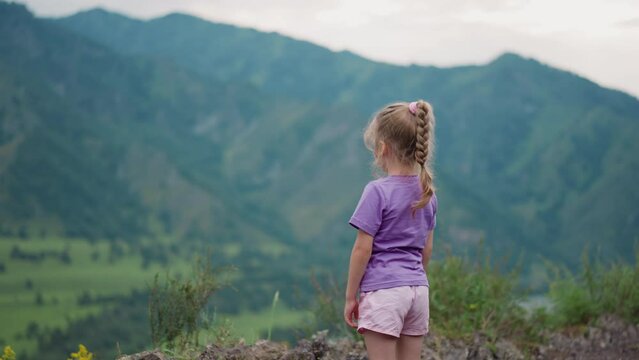 Dreaming little girl in lilac t-shirt scratches leg with mosquito bite on hill top edge against mountains on gloomy day backside view slow motion