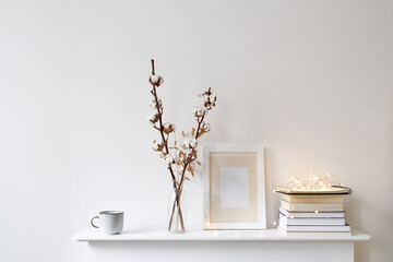 Scandinavian style room interior in white tones. A vase with cotton flowers, a stack of books, a photo frame, a garland on a wooden surface of a shelf. Copy space.