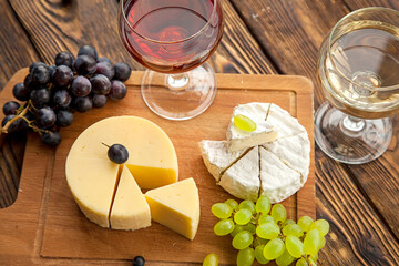 Various types of cheese with dark and green grapes and wine on wooden table.