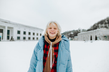 Portrait of a joyful young girl in a warm winter jacket on a walk on a snowy street background, looking at the camera and smiling.