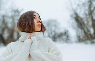 Attractive woman in a knitted sweater stands on the street in winter and looks away with a serious face on a background of snowy views.