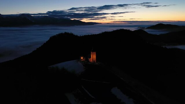 Illuminated Church of St. Primoz and Felicijan at Dawn above the Clouds. Julian Alps. Jamnik, Slovenia, Europe. Aerial View. Orbiting