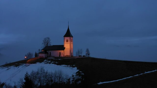 Illuminated Church of St. Primoz and Felicijan at Winter Evening Foggy Twilight. Cloudy Sky. Jamnik, Slovenia. Aerial View. Orbiting