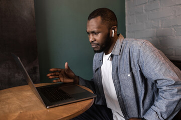 Pensive young African American businessman looks at laptop screen, thinks over a plan or strategy of an online project, solves problems, student works on a difficult task, does homework.