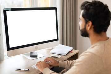 Back view of positive Hindi man in smart casual clothes using computer while sitting at the desk in his flat. Young Indian male student watching webinars, educational courses, learning on the distance