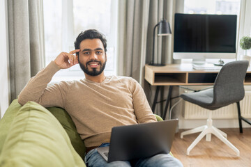 Front view of positive Hindi man in smart casual clothes using laptop while sitting at the sofa in...