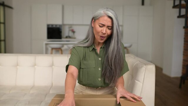 Satisfied stylish senior Asian woman opens parcel carton box, unpacking received gift, feeling curious about ordered item from an online store, female customer happy with fast delivery and shipment
