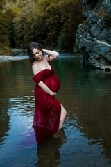 Young, elegant, blonde pregnant woman standing in the river in red dress
