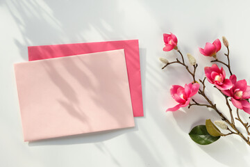 Blank postcard with envelope. Pink decorative magnolia flowers. Top view on off white background. Natural direct sunlight, long shadows.