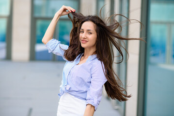 Cheerful longhair girl revolve around relaxing in city downtown. Carefree brunette woman enjoy walking outdoors on weekend. Cute asian tourist posing touching beautiful hair in front high buildings.