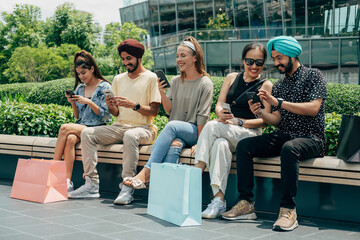 Group Of Five Happy Multi Ethnic Friends Using Mobile Phones Outdoors. 
Smiling friends watching something on the phone, talking and laughing while sitting in a row on a bench with shopping bags.