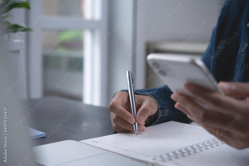 Wall mural young business woman sitting at table, writing notes on notebook, using mobile phone and working on 