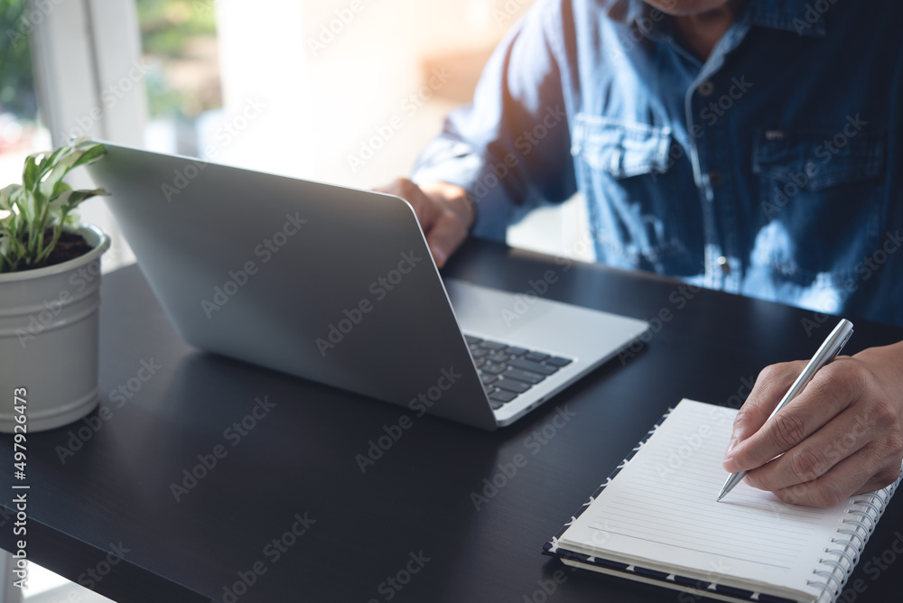 Poster close up of man working on laptop computer and hand with a pen writing on notebook on office table