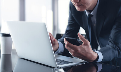 Asian businessman in black suit busy working on laptop computer with mobile phone and digital tablet on table at office, close up.