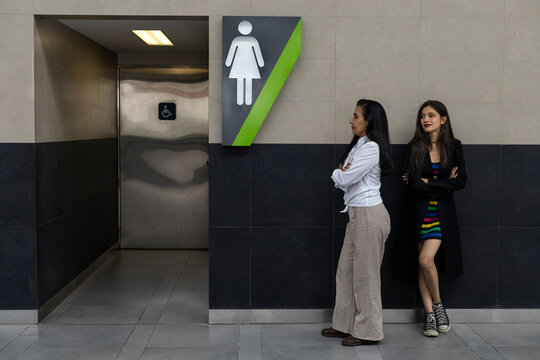 Two adult Latin American women mother and daughter urgently waiting in line to enter public restroom. Sharing concept