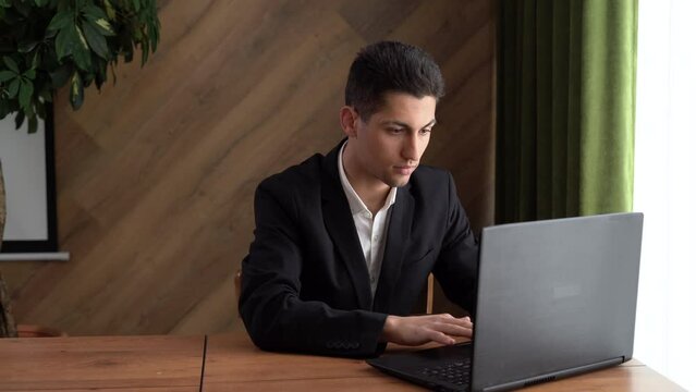 Young arabian male using laptop computer, sitting at table in home office, typing on keyboard and looking at screen, enjoying remote work