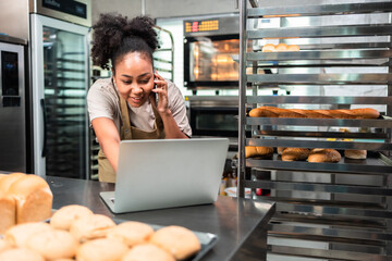 Young  African female in apron using laptop and talking to clients on the phone by workplace.Woman...