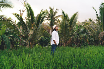 Balinese male farmer in casual clothes spending daytime at rice fields during travel vacations in Indonesia, adult businessman in hat visiting own countryside plantation with paddy in Thailand