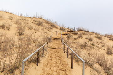 Fototapeta na wymiar The staircase rises to a sandy dune. Climbing to the top in the desert. Sarykum dune. Dagestan, Russia. A unique sandy mountain in the Caucasus on a cloudy day. Grass grows on a sand dune.