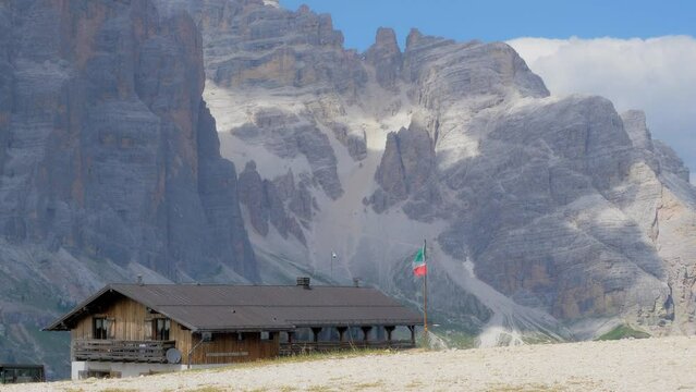 High mountain refuge with an Italian flag that waves in the wind in a summer day is surrounded by still snowy mountains