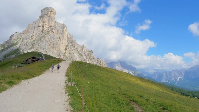 Couple of hikers walk on a sand and stone pathway in high mountain that pass thru a meadow, with their backpack and the specific mountain equipment, to reach the summit of a rock in a summer day