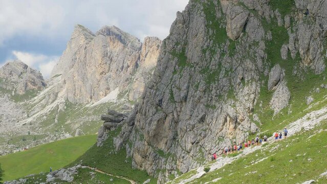 A group of trekkers walk in a row on a high mountain path that winds through the green grass. They all wear sports gear. All around bare rock mountains stand out on a sunny day
