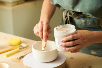 Close-up of girl painting clay mug with glaze. Woman coloring pottery in workshop with a paintbrush. Painter in green apron glazing clay pot.