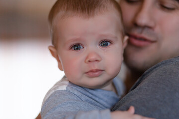 Closeup Sad young blond boy crying on father hands in room. Man holds son, hugs and comforts. Family love, care and moral support, baby's tears, daddy's arms. Dad consoling crying child