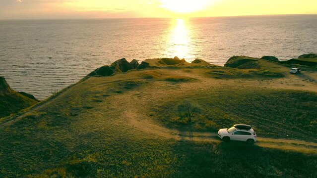 Scenic hilly coastline with suv on dirty road with dramatic marine sunset on background. Aerial view from drone of white car riding country road, turning to sea coast at dusk. Concept of destination
