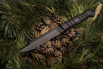 Tactical knife on a Christmas tree background. Black knife, shot against the background of green branches of a Christmas tree - obrazy, fototapety, plakaty
