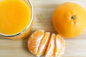 Top view of glass of natural orange juice in foreground, peeled orange pieces and full orange in background, on wooden surface, selective focus used