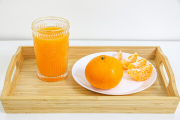Naklejka premium Glass of sweet and natural orange juice, full of vitamin goodness, besides full round orange and peeled orange pieces in a wooden tray against white background