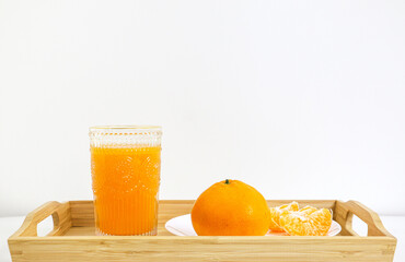 Naklejka premium A healthy glass of natural orange juice besides a full orange and peeled orange pieces in a wooden tray against white background
