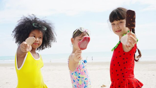 Group of Little African and Asian girl eating ice cream together while playing on tropical beach together in sunny day. Happy children family enjoy and fun outdoor activity on summer holiday vacation