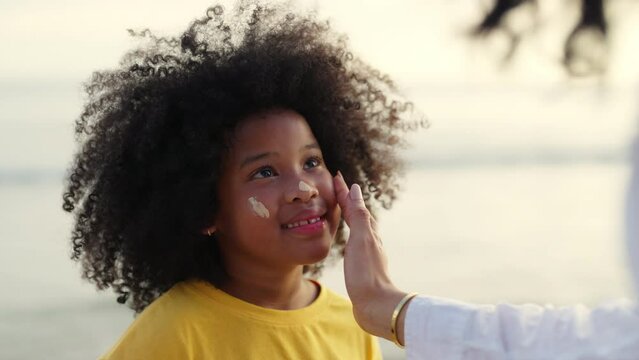 Happy African family mom and child girl kid enjoy and fun outdoor lifestyle on beach vacation. Mother applying sunblock to little daughter while walking together on tropical beach at summer sunset.